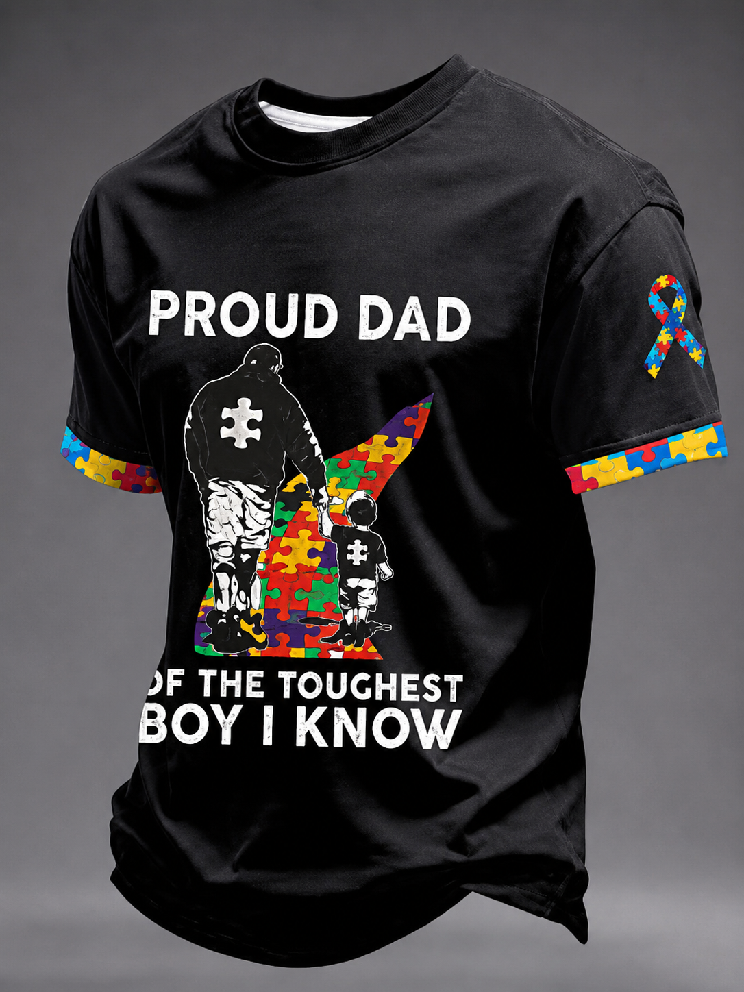 A black short-sleeve graphic T-shirt displayed on a gray background. The shirt features a large front print with the words 'PROUD DAD' and 'OF THE TOUGHEST BOY I KNOW' in white, alongside a silhouette of an adult and child made from multicolored puzzle pieces.