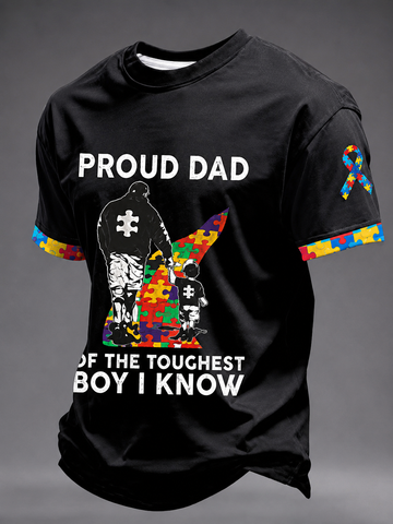 A black short-sleeve graphic T-shirt displayed on a gray background. The shirt features a large front print with the words 'PROUD DAD' and 'OF THE TOUGHEST BOY I KNOW' in white, alongside a silhouette of an adult and child made from multicolored puzzle pieces.