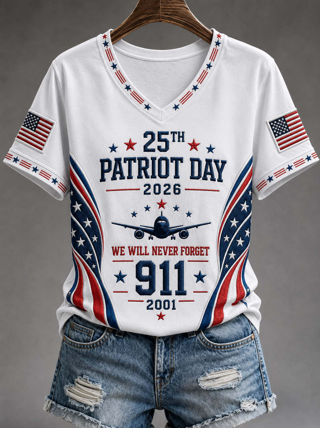 White V-neck T-shirt with patriotic red, white, and blue graphics celebrating 25th Patriot Day 2026, featuring an airplane, stars, and American flag sleeve prints, shown on a hanger with distressed denim shorts against a gray background.