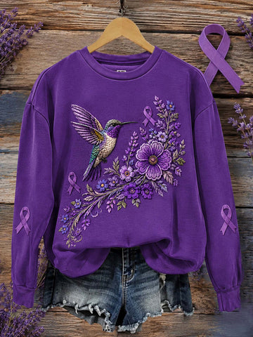 A purple crewneck sweatshirt with a detailed graphic of a hummingbird and purple flowers, styled on a wooden hanger with denim shorts against a rustic wood plank background.