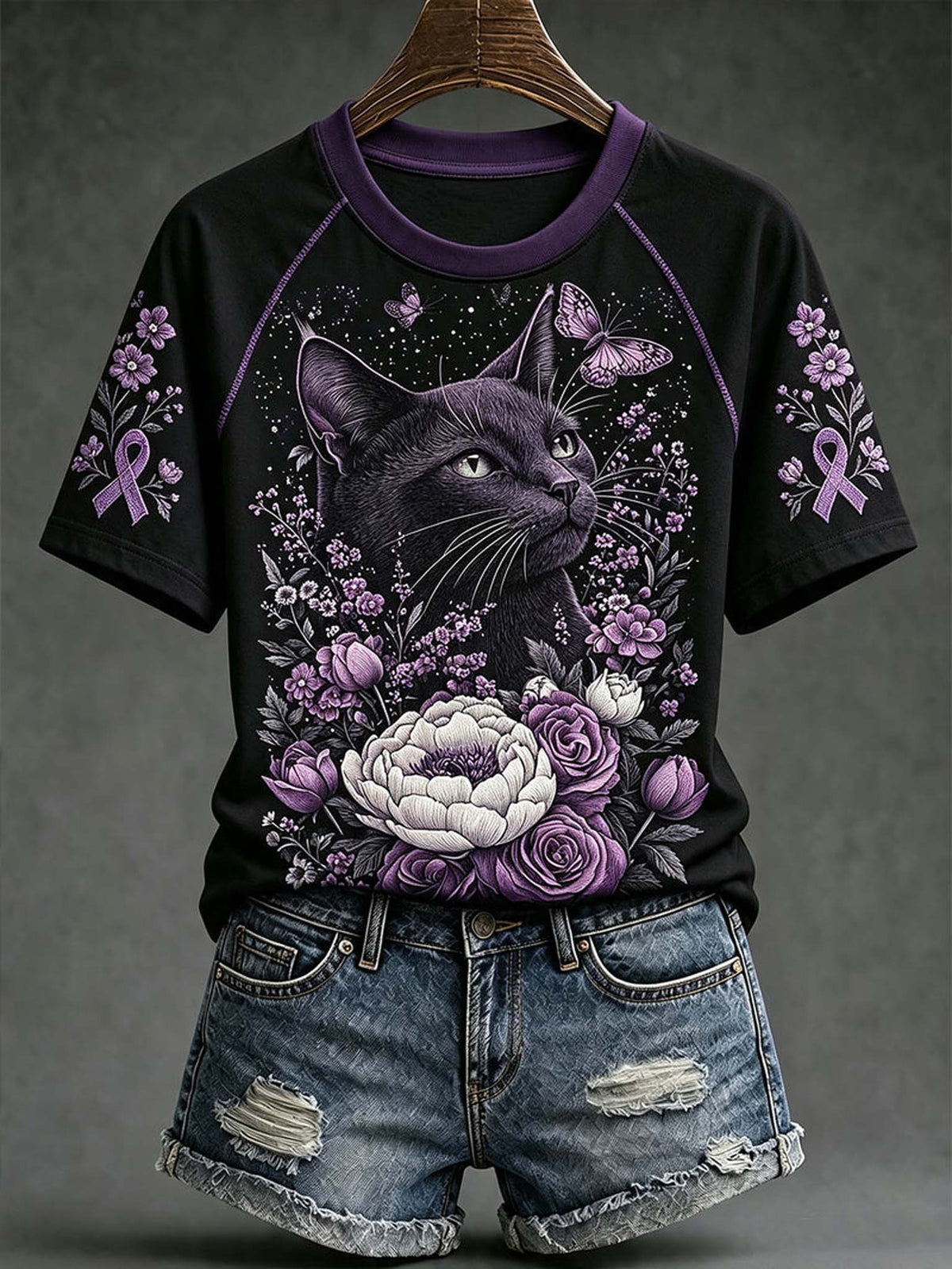 A black short-sleeve raglan T-shirt with a purple and black cat graphic surrounded by purple flowers, ribbons, and butterflies, styled with distressed denim shorts on a hanger against a dark gray background.