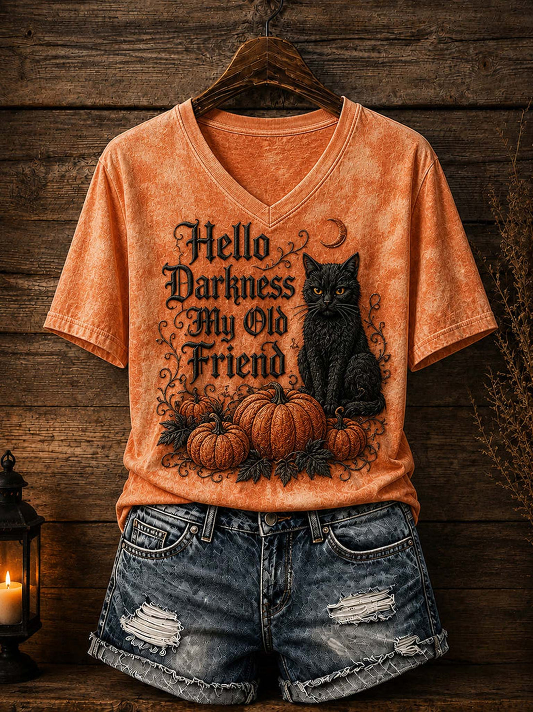 An orange acid-wash V-neck t-shirt with a Halloween graphic reading 'Hello Darkness My Old Friend' alongside a black cat and pumpkins, styled on a wooden hanger with distressed denim shorts against a rustic wood plank background.