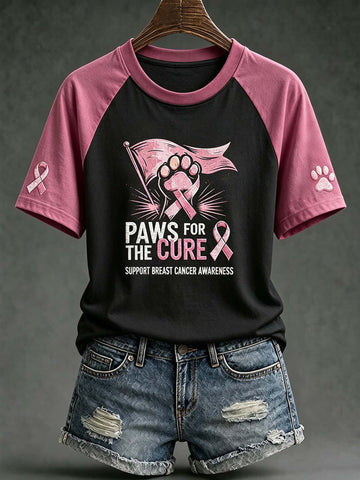 A black and pink raglan t-shirt with a 'Paws for the Cure' breast cancer awareness graphic, styled on a wooden hanger with distressed denim shorts against a dark gray background.