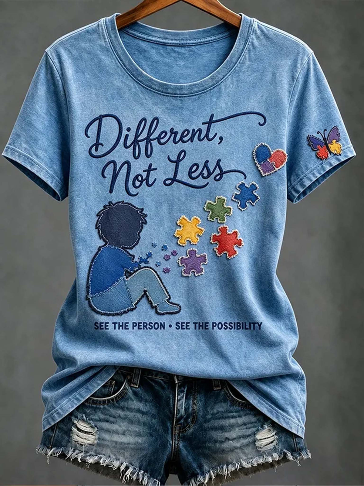 A light blue graphic t-shirt on a wooden hanger, styled with distressed denim shorts against a neutral gray background. The t-shirt features a graphic of a child sitting with colorful puzzle pieces and the text 'Different, Not Less' and 'SEE THE PERSON * SEE THE POSSIBILITY'.