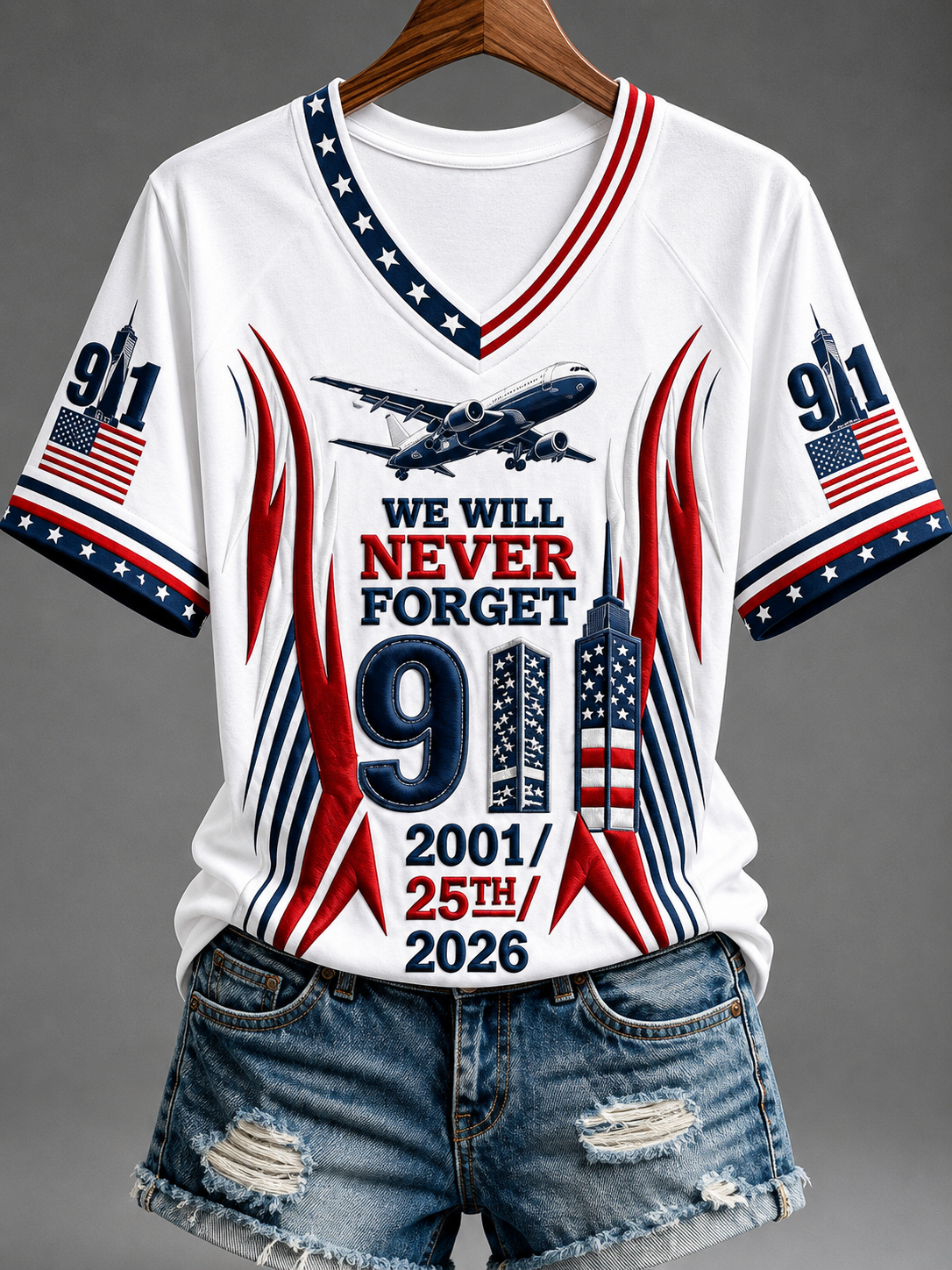 A white V-neck t-shirt with a patriotic 9/11 memorial graphic, styled on a wooden hanger with distressed denim shorts against a gray background.