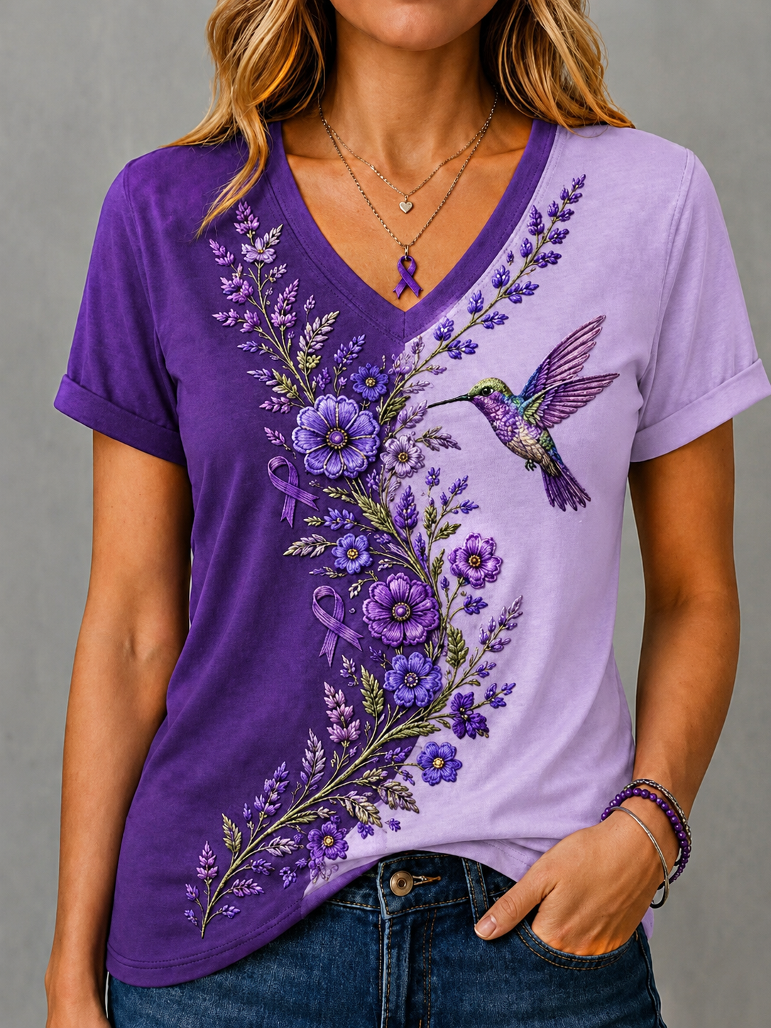 A woman wearing a purple and lavender V-neck t-shirt with a floral and hummingbird graphic, paired with blue jeans, against a neutral gray background.