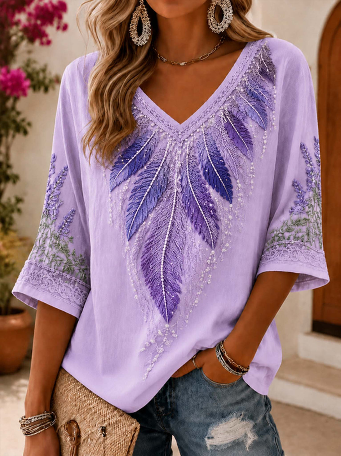 A woman wearing a lavender V-neck top with purple feather embroidery and beadwork, paired with distressed blue jeans and gold jewelry, in an outdoor setting.