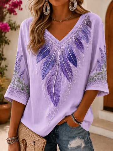 A woman wearing a lavender V-neck top with purple feather embroidery and beadwork, paired with distressed blue jeans and gold jewelry, in an outdoor setting.