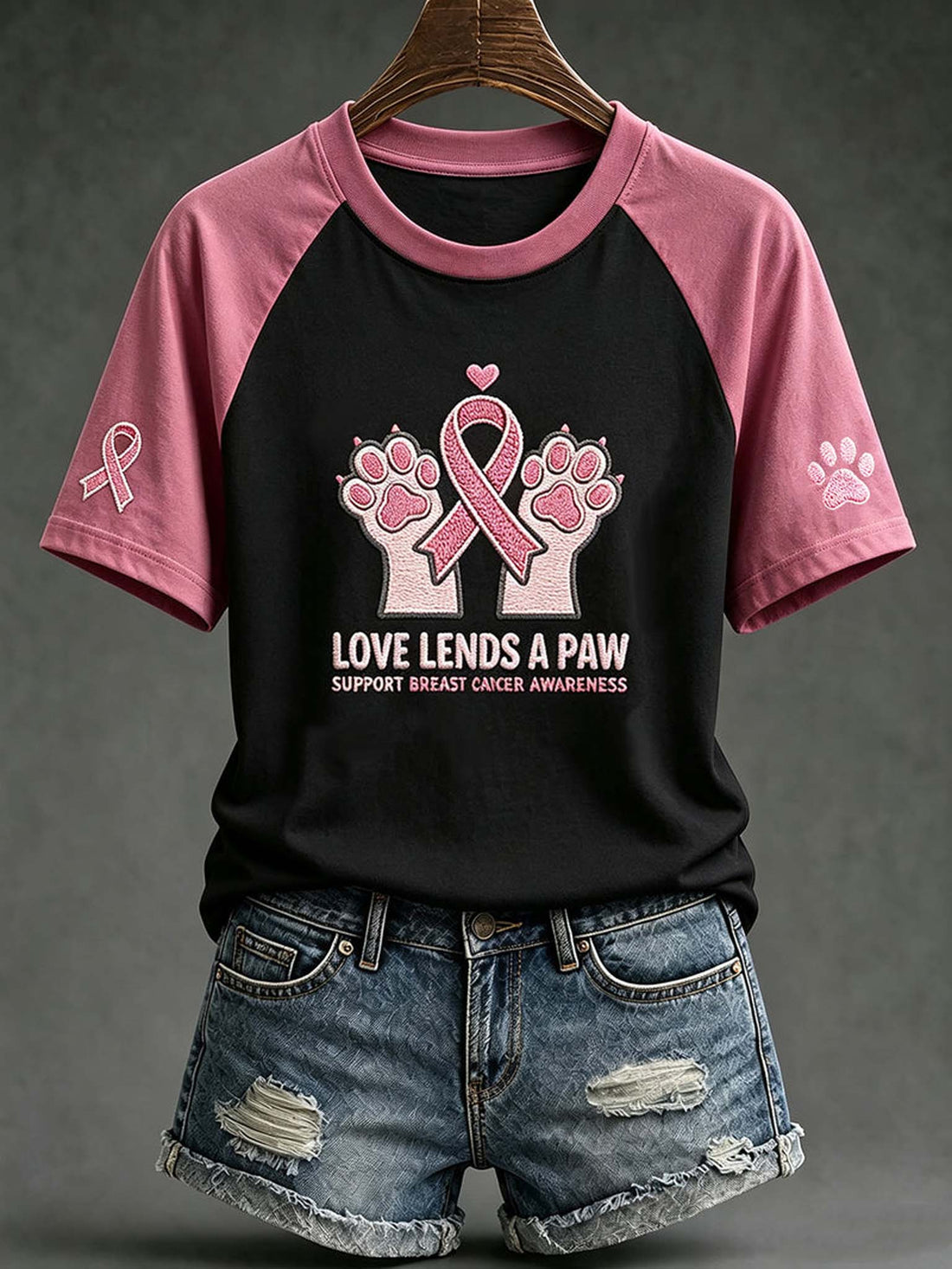A women's short-sleeve raglan T-shirt hanging on a wooden hanger, paired with distressed denim shorts. The shirt has a black body and pink sleeves, featuring a graphic of two paw prints with a pink ribbon and the text 'LOVE LENDS A PAW SUPPORT BREAST CANCER AWARENESS' on the front.