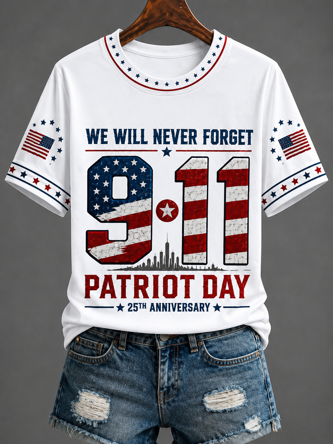 White short-sleeve graphic T-shirt on a hanger with distressed blue denim shorts below. The shirt features a patriotic 9/11 memorial design with the text 'WE WILL NEVER FORGET 9/11 PATRIOT DAY 25TH ANNIVERSARY' and American flag motifs.