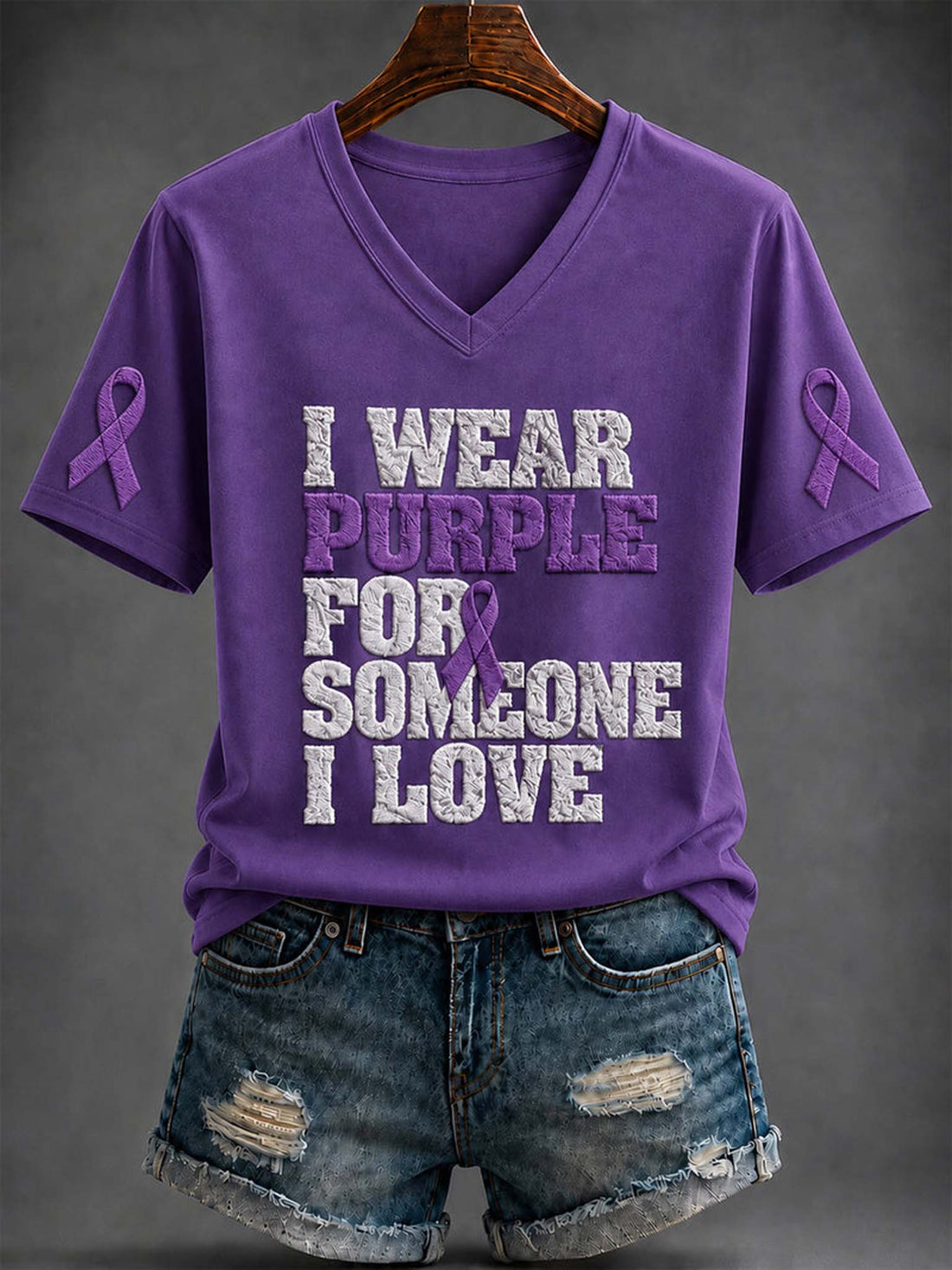 A purple V-neck T-shirt on a wooden hanger, styled with distressed denim shorts against a dark gray background. The shirt features the text 'I WEAR PURPLE FOR SOMEONE I LOVE' in distressed white and purple lettering, with purple awareness ribbons on both sleeves.
