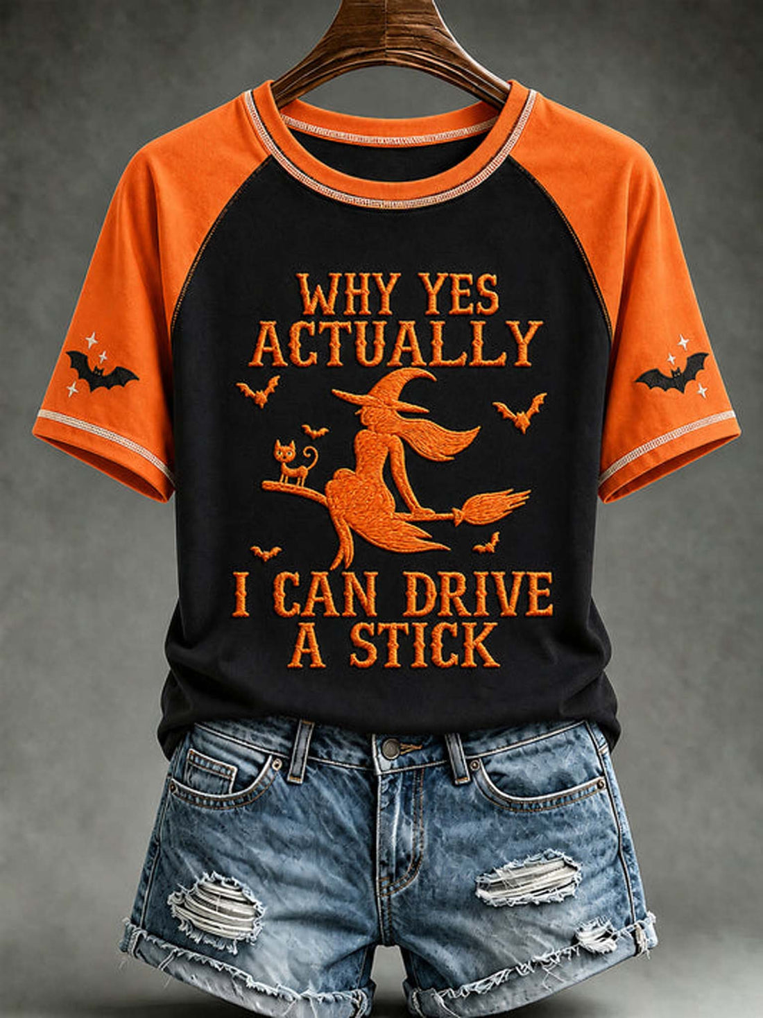 A raglan-style short-sleeve T-shirt with bright orange sleeves and collar trim, paired with distressed denim shorts. The shirt has a black front panel featuring an orange witch riding a broomstick with the text 'WHY YES ACTUALLY I CAN DRIVE A STICK' and small bat motifs.