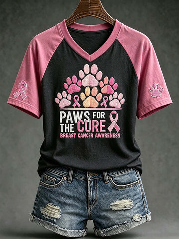 A women's short-sleeve raglan V-neck T-shirt on a hanger, featuring a black body and pink sleeves. The front has a graphic of pink, lavender, and peach paw prints above the text 'PAWS FOR THE CURE BREAST CANCER AWARENESS' with pink ribbons.