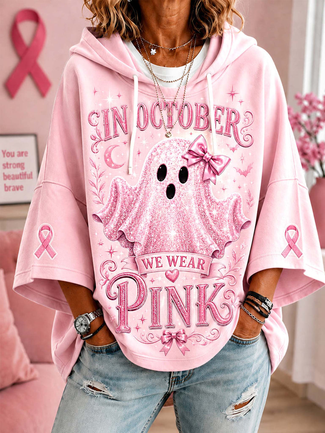 A woman wearing a pink graphic hoodie with a ghost design and the text 'IN OCTOBER WE WEAR PINK', paired with light-wash distressed jeans in a pink room setting.