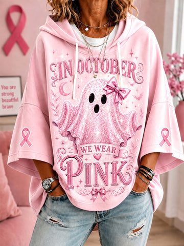 A woman wearing a pink graphic hoodie with a ghost design and the text 'IN OCTOBER WE WEAR PINK', paired with light-wash distressed jeans in a pink room setting.
