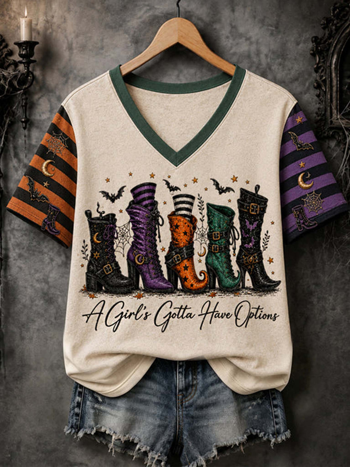 A cream-colored v-neck t-shirt with green trim, featuring a graphic of four colorful witch boots and the text 'A Girl's Gotta Have Options' on the front. The sleeves have a black and orange striped pattern on the left and a purple pattern on the right, shown hanging on a wooden hanger with denim shorts against a dark textured background.