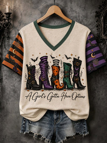A cream-colored v-neck t-shirt with green trim, featuring a graphic of four colorful witch boots and the text 'A Girl's Gotta Have Options' on the front. The sleeves have a black and orange striped pattern on the left and a purple pattern on the right, shown hanging on a wooden hanger with denim shorts against a dark textured background.