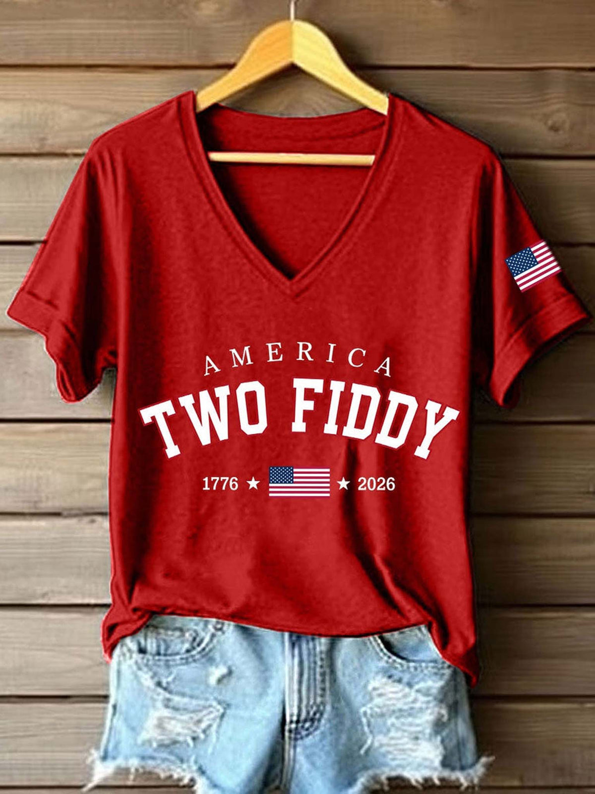 A red V-neck t-shirt with 'AMERICA TWO FIDDY 1776 2026' printed in white on the front, styled on a wooden hanger with distressed denim shorts against a wooden plank wall. The t-shirt also features a small American flag graphic on the right sleeve.