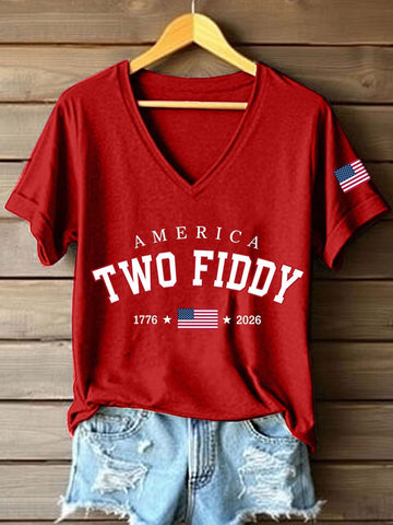 A red V-neck t-shirt with 'AMERICA TWO FIDDY 1776 2026' printed in white on the front, styled on a wooden hanger with distressed denim shorts against a wooden plank wall. The t-shirt also features a small American flag graphic on the right sleeve.