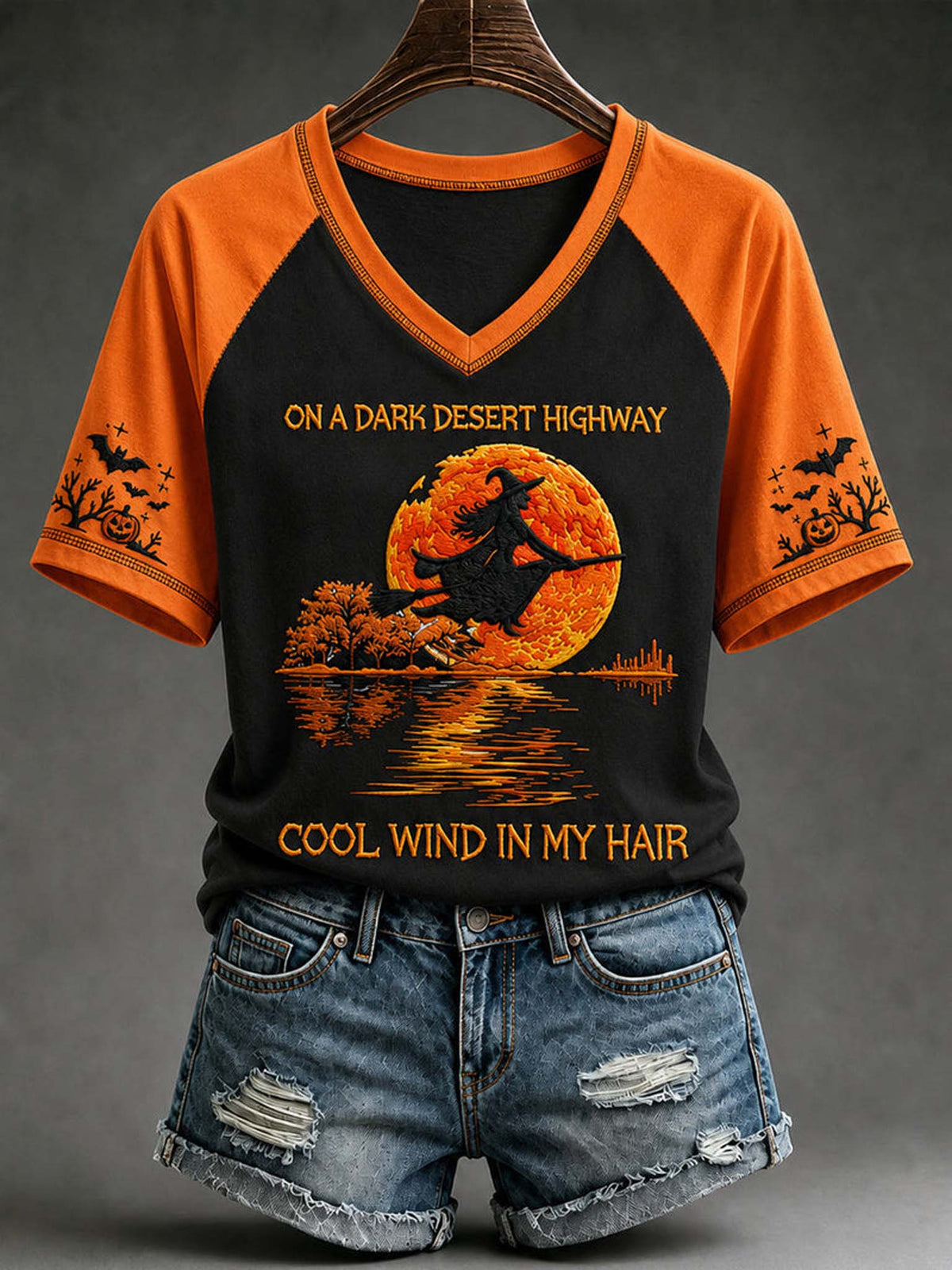 A black and orange raglan V-neck t-shirt with a Halloween graphic of a witch riding a broomstick in front of a full moon, styled on a hanger with distressed denim shorts against a dark gray background.