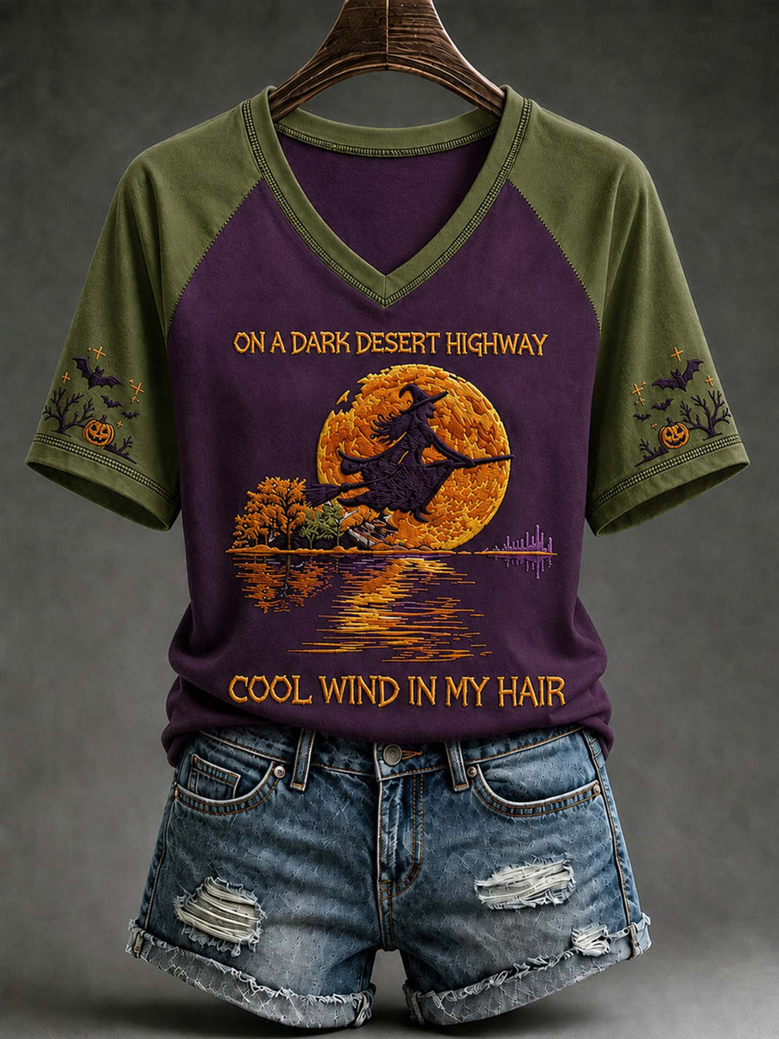 A raglan-style V-neck T-shirt with olive green sleeves and collar, and a dark purple body, featuring a Halloween graphic of a witch riding a broomstick across a large orange full moon over water. The text reads 'ON A DARK DESERT HIGHWAY' and 'COOL WIND IN MY HAIR.
