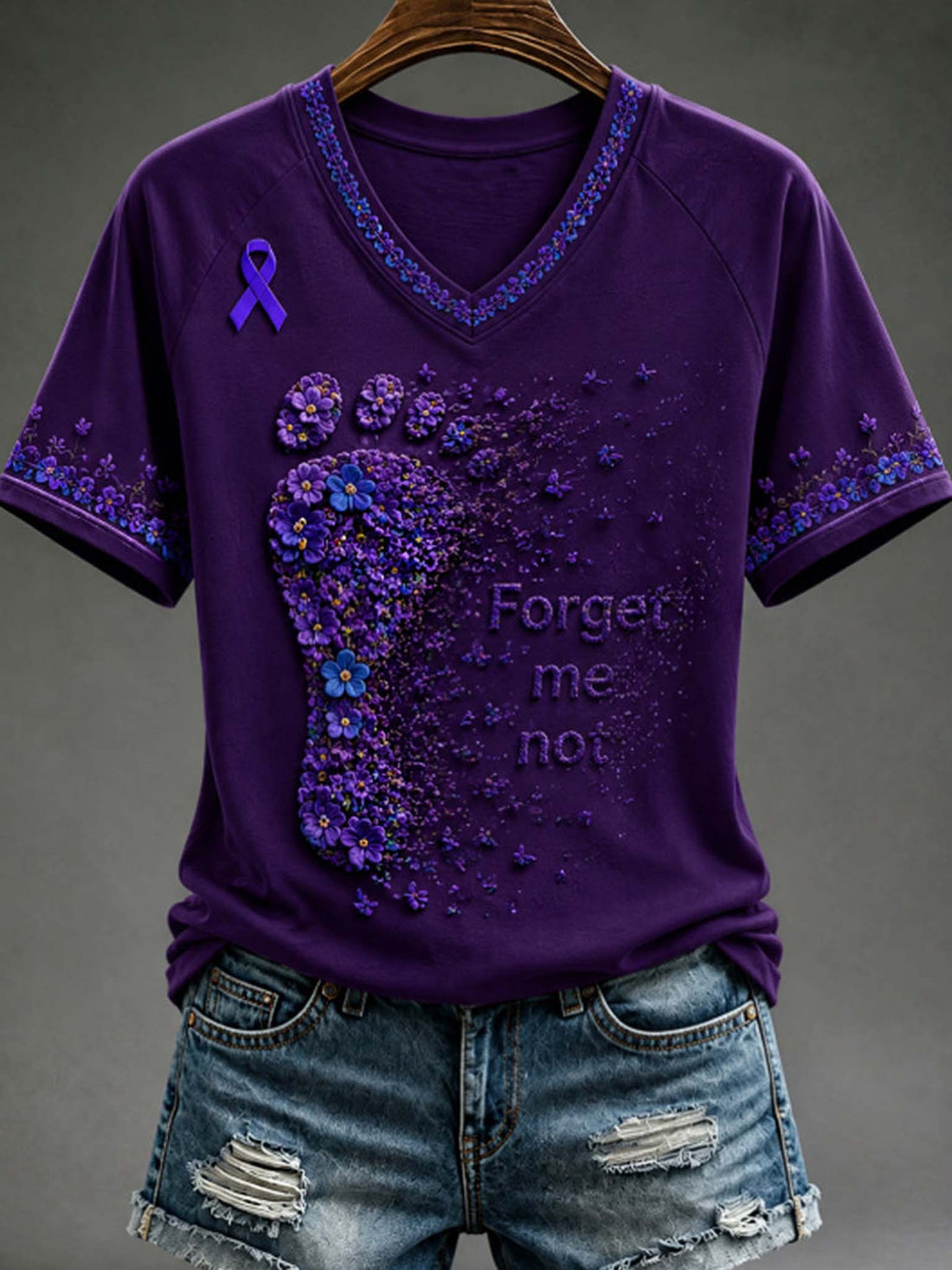 A purple short-sleeve V-neck T-shirt with a large purple foot-shaped floral design, a purple awareness ribbon on the left chest, and the words 'Forget me not' printed on the front, shown hanging on a wooden hanger with distressed denim shorts against a dark gray background.