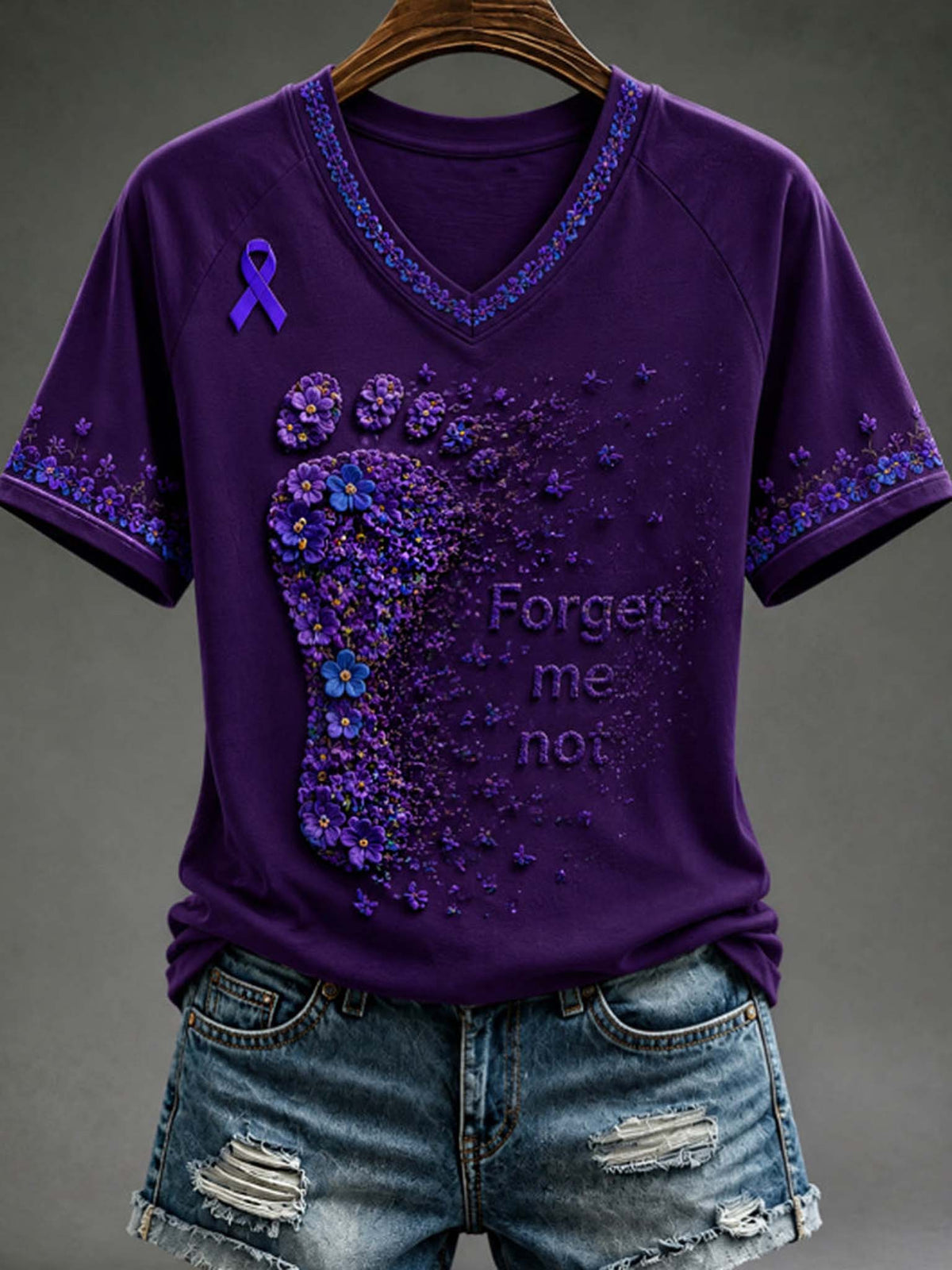 A purple short-sleeve V-neck T-shirt with a large purple foot-shaped floral design, a purple awareness ribbon on the left chest, and the words 'Forget me not' printed on the front, shown hanging on a wooden hanger with distressed denim shorts against a dark gray background.