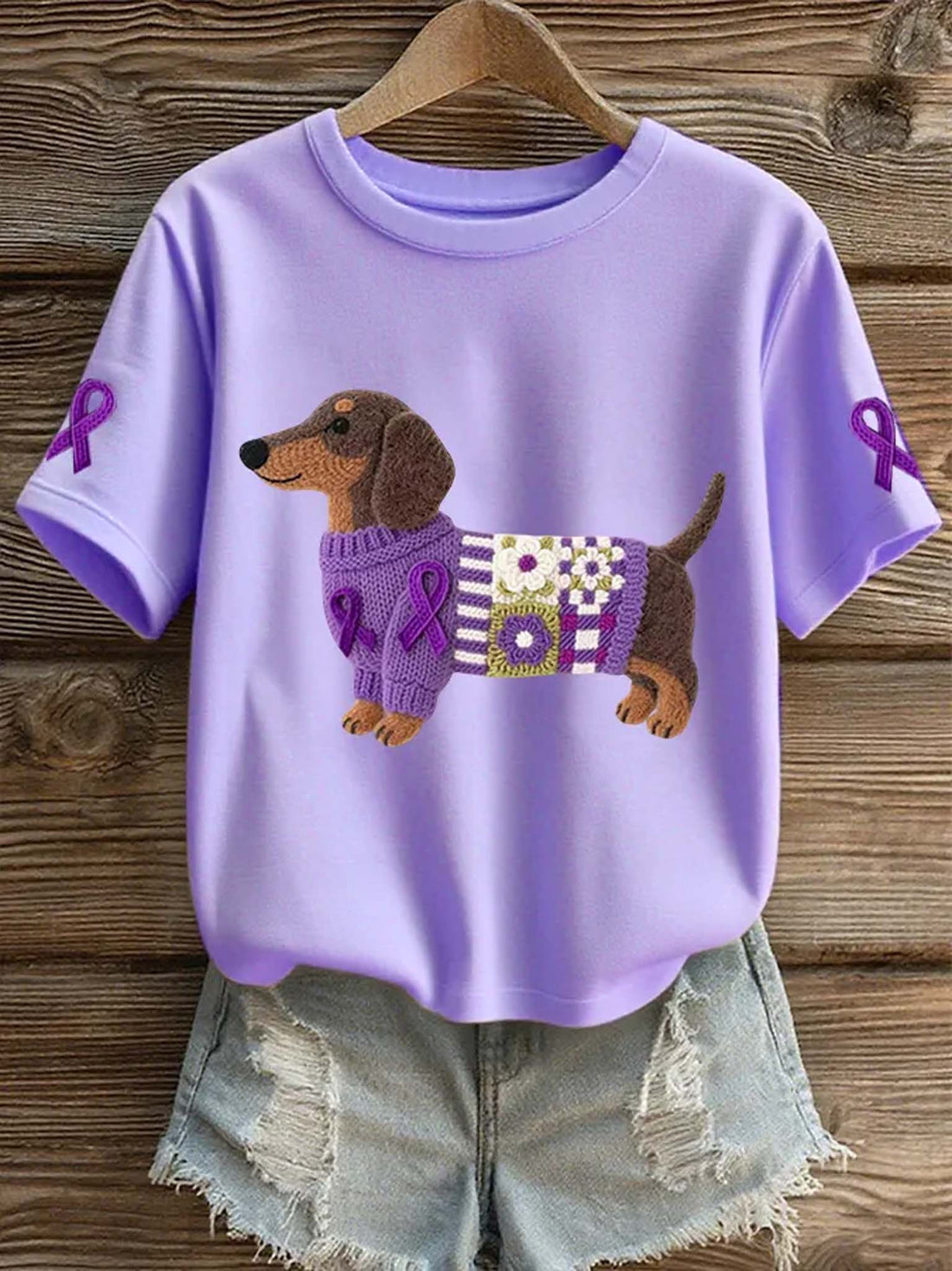 A lavender purple short-sleeve t-shirt with a dachshund wearing a knitted purple sweater, styled with distressed denim shorts on a wooden hanger against a rustic wood plank background.