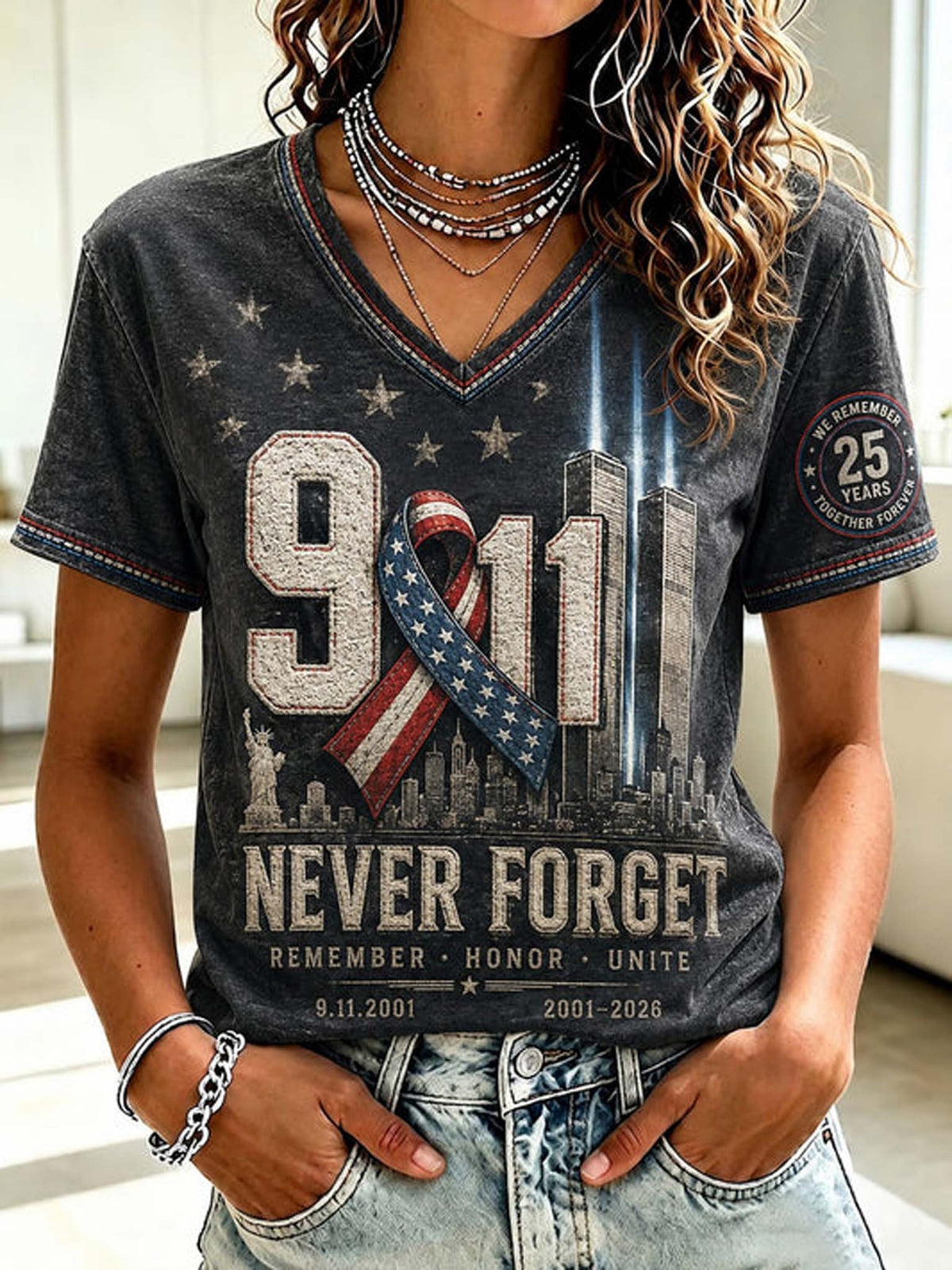 A woman wearing a dark gray V-neck graphic t-shirt with a '9/11 Never Forget' design featuring the New York City skyline, a patriotic ribbon, and 'Remember Honor Unite' text, paired with light wash denim jeans.