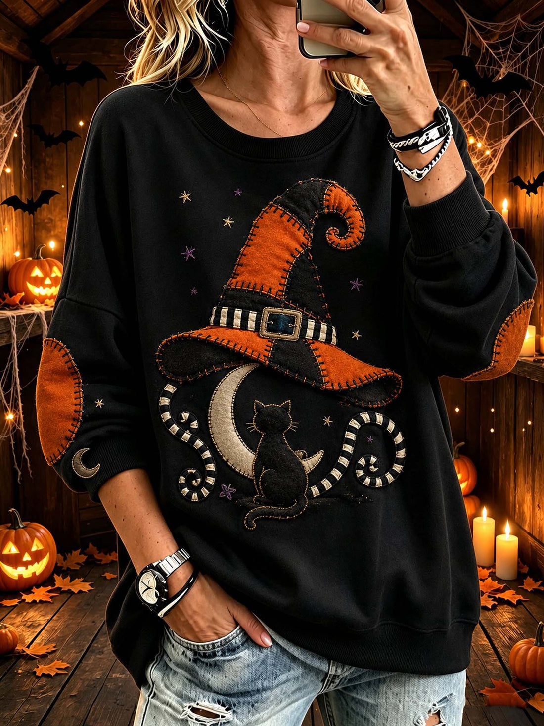 A woman wearing a black long-sleeve sweatshirt with a large Halloween-themed patchwork design featuring an orange and black witch hat, a black cat, a crescent moon, and stars. She is taking a mirror selfie in a cozy, warmly lit Halloween-themed room decorated with pumpkins, candles, and cobwebs.