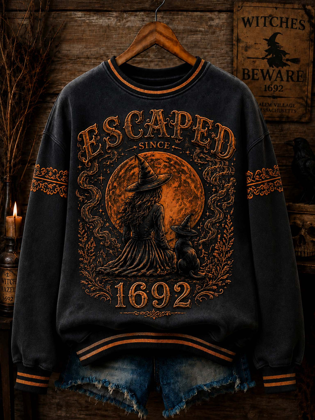 A dark charcoal gray crewneck sweatshirt with a distressed orange-brown Halloween graphic featuring a witch, cat, full moon, and the text 'ESCAPED SINCE 1692'. The sweatshirt is styled on a wooden hanger with frayed blue denim shorts against a rustic wooden background with a 'Witches Beware' sign.