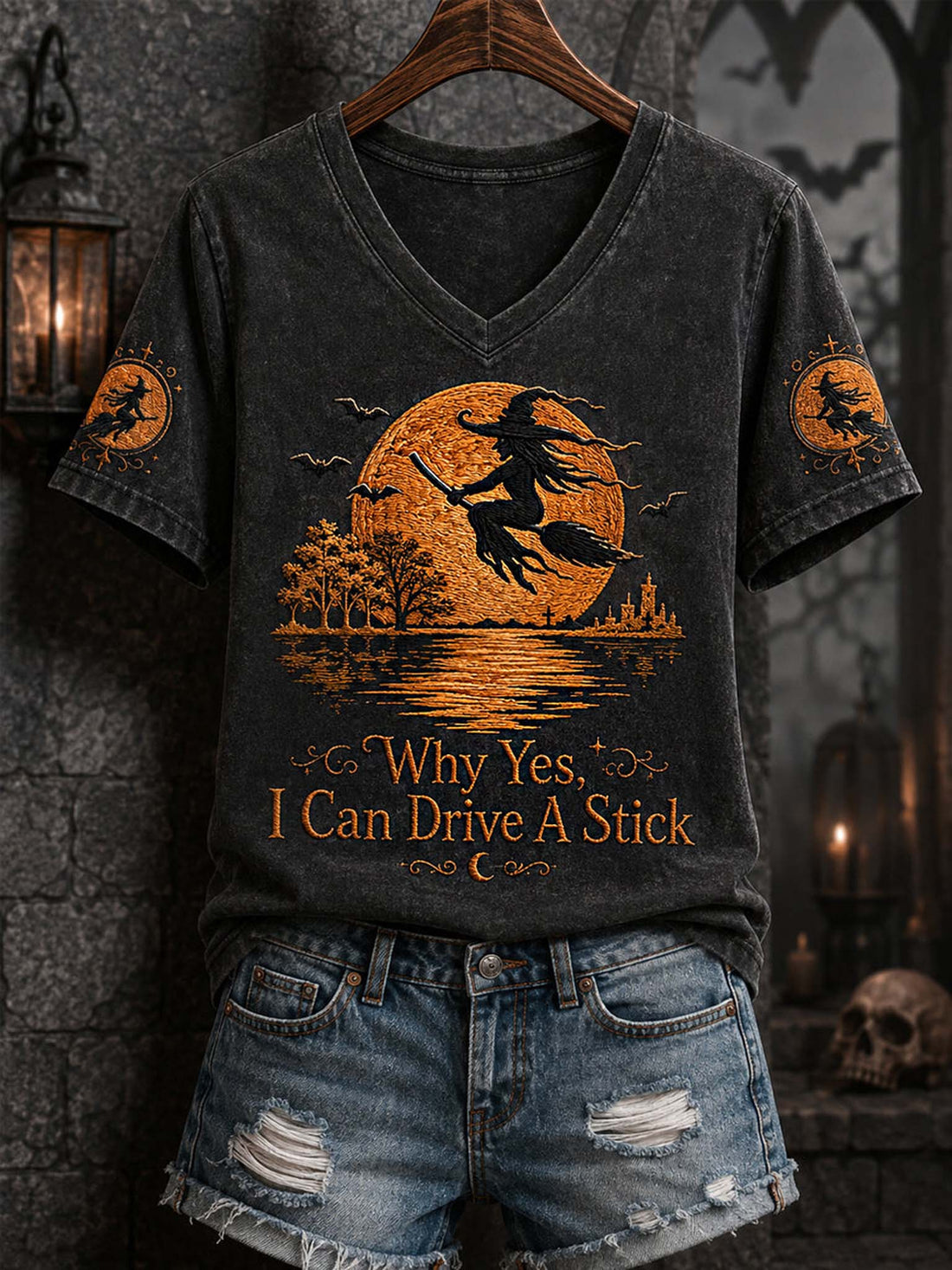 A dark gray washed V-neck t-shirt with a Halloween graphic of a witch riding a broomstick in front of a full moon, displayed on a wooden hanger with distressed denim shorts against a dark gothic background.