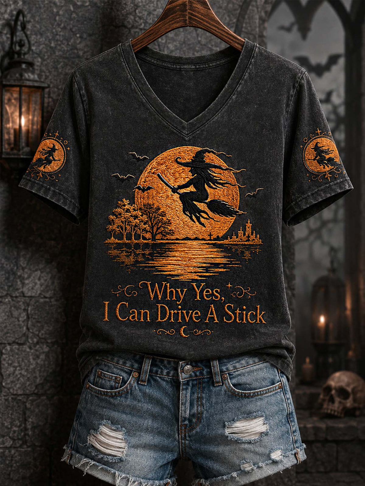A dark gray washed V-neck t-shirt with a Halloween graphic of a witch riding a broomstick in front of a full moon, displayed on a wooden hanger with distressed denim shorts against a dark gothic background.
