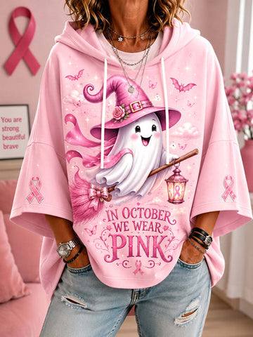 A woman wearing a pink hoodie with a cartoon ghost wearing a witch hat, holding a lantern, and the text 'IN OCTOBER WE WEAR PINK' on the front. The hoodie also features pink ribbon graphics on the sleeves.