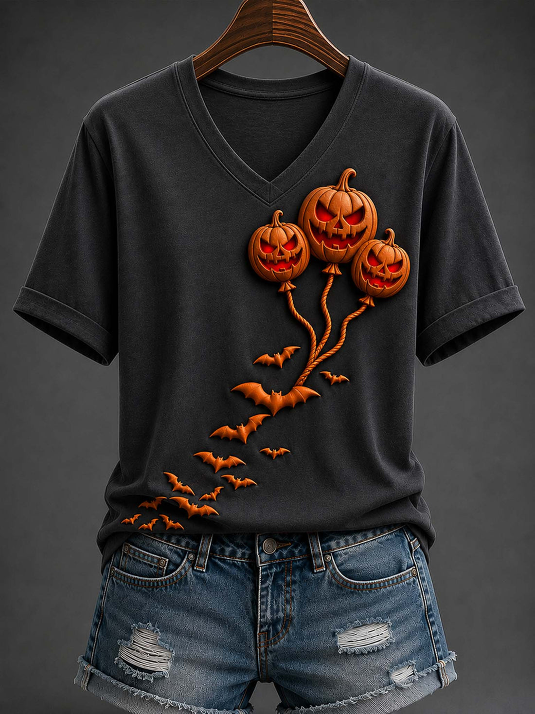 A dark charcoal gray V-neck t-shirt with a 3D-effect graphic of three glowing jack-o'-lantern balloons and flying bats, styled on a wooden hanger with distressed denim shorts against a dark gray background.