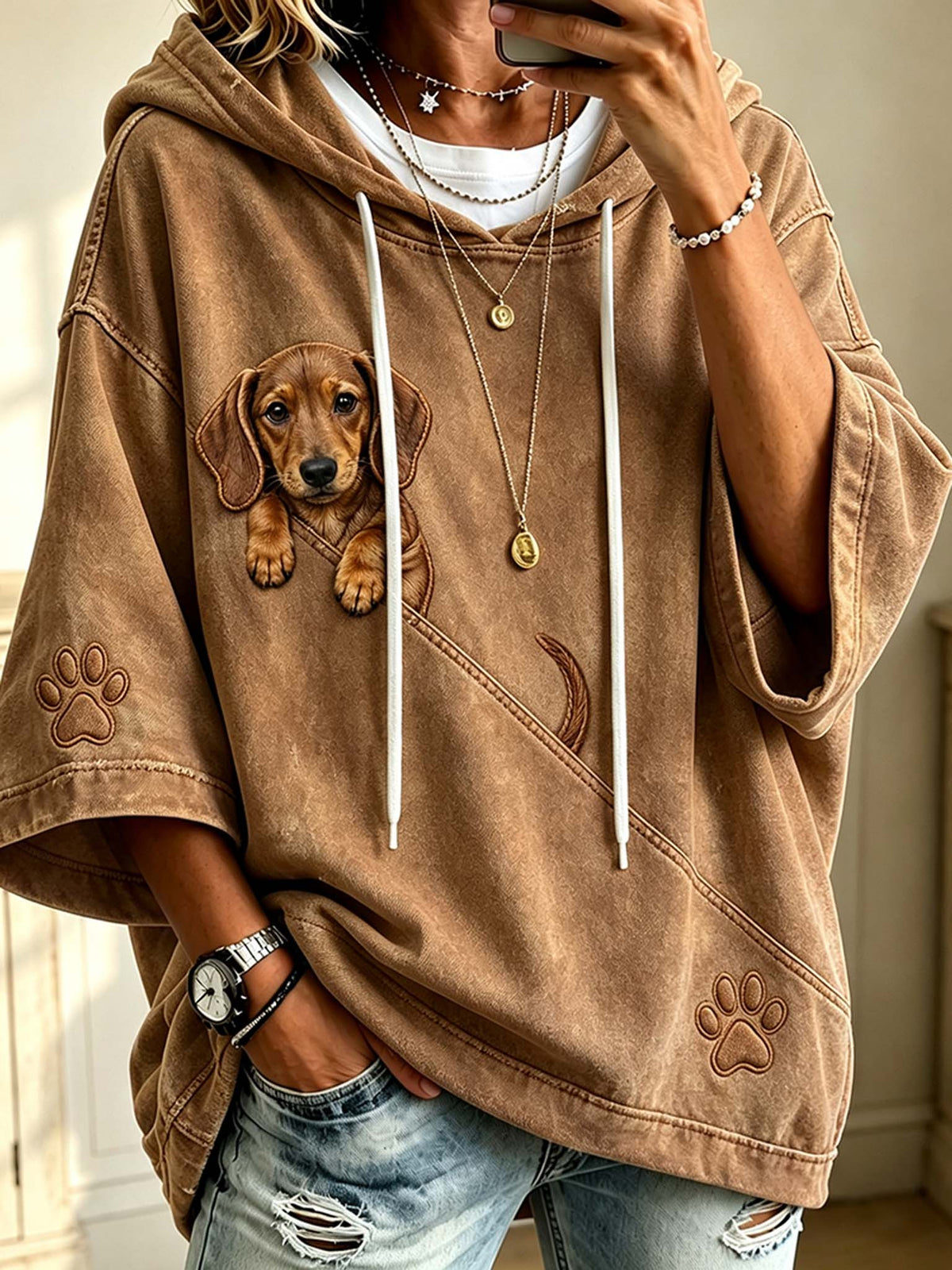 A person wearing a brown oversized hoodie with a dachshund graphic on the front, white drawstrings, and paw print details on the sleeves, paired with distressed blue jeans.