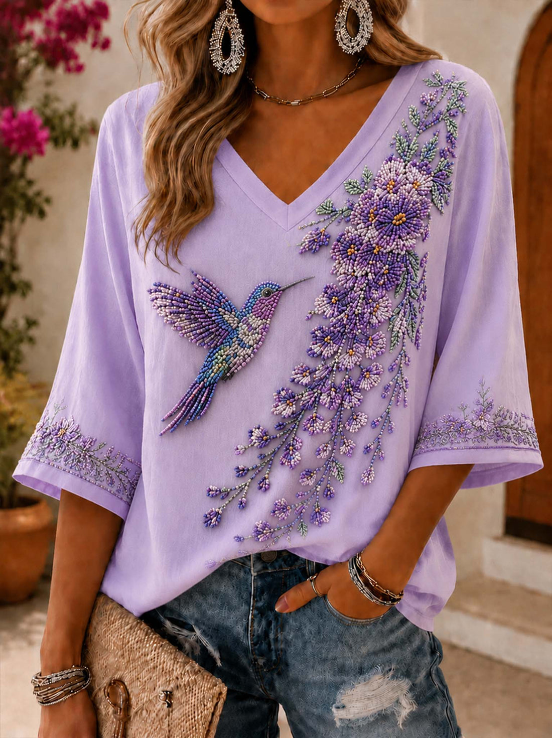 A woman wearing a lavender V-neck top with intricate beaded floral and hummingbird embroidery, paired with distressed blue jeans and holding a woven clutch.