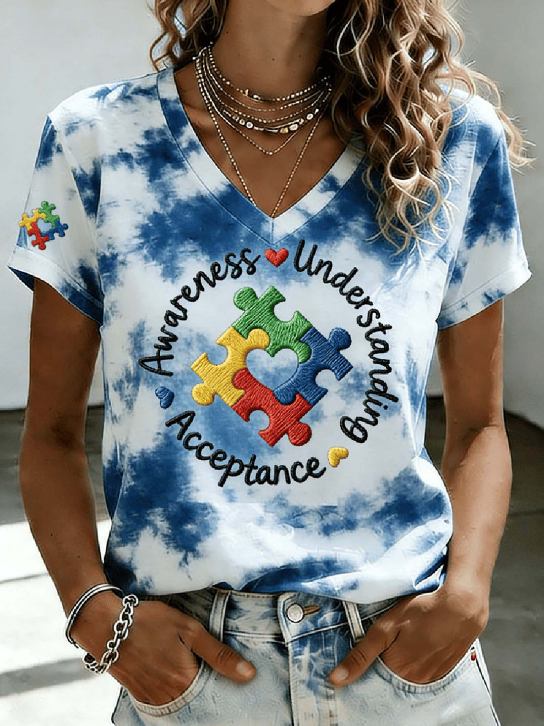 A woman wearing a blue and white tie-dye V-neck t-shirt with a colorful puzzle piece graphic and the words 'Awareness Understand Acceptance' on the front, paired with light-wash denim jeans.