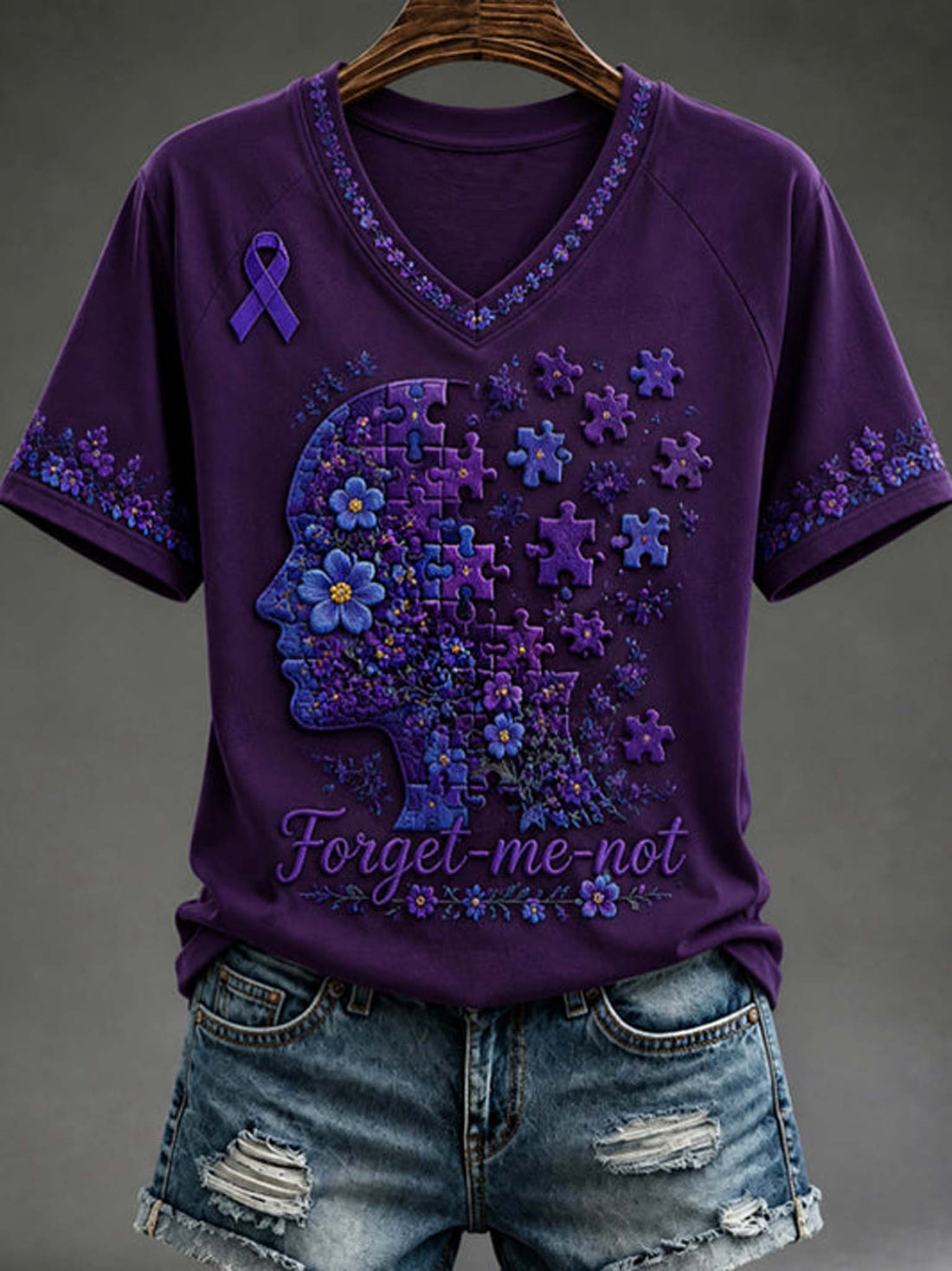 A purple short-sleeve V-neck T-shirt with a large floral and puzzle-piece head graphic in shades of purple and blue, a purple awareness ribbon on the left chest, and the words 'Forget-me-not' across the lower front. The shirt is displayed on a hanger above distressed denim shorts against a dark gray studio background.
