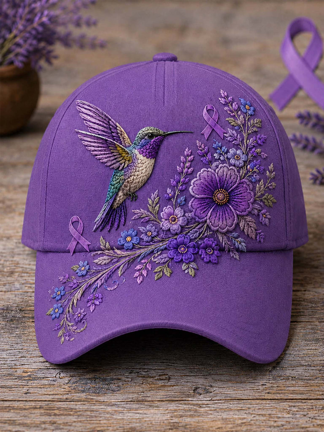 A purple baseball cap with detailed embroidery of a hummingbird and purple flowers, ribbon motifs, and floral branches, displayed on a rustic wooden surface.