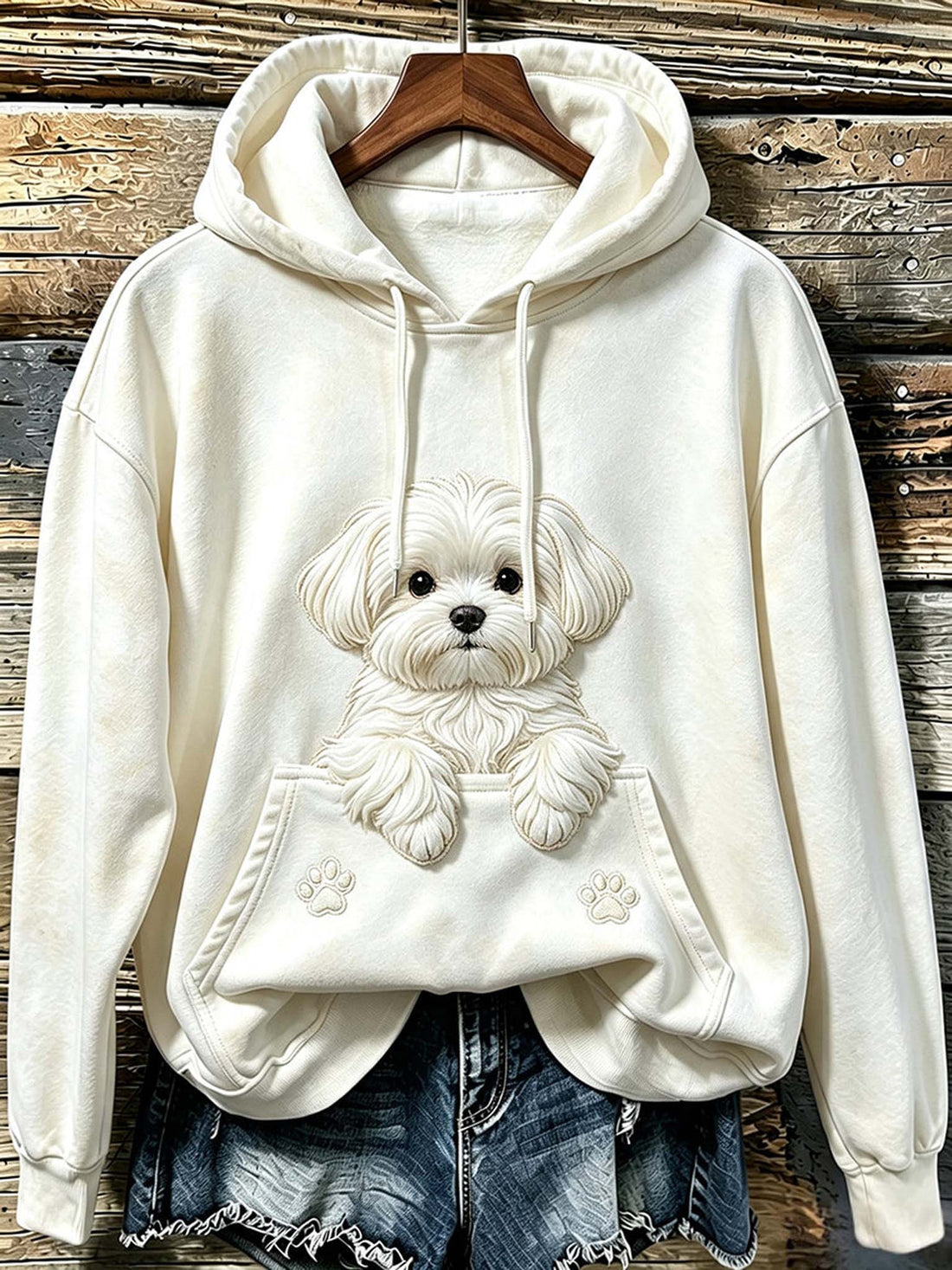 Cream-colored hoodie with a 3D-effect embroidered dog design on the front, styled with denim shorts on a wooden hanger against a rustic wood plank background.