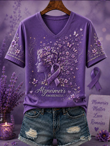 A purple short-sleeve V-neck T-shirt displayed on a hanger with distressed denim shorts, featuring a lavender awareness ribbon, floral design, and the words 'Alzheimer's AWARENESS' on the front. The shirt is styled with lavender stems, a candle, and a memorial sign against a purple background.
