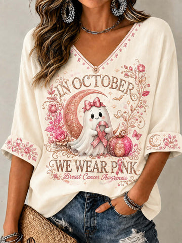 A woman wearing a cream-colored V-neck t-shirt with a pink and white graphic of a cute ghost holding a pink breast cancer awareness ribbon, surrounded by the text 'IN OCTOBER WE WEAR PINK' and 'Breast Cancer Awareness'.