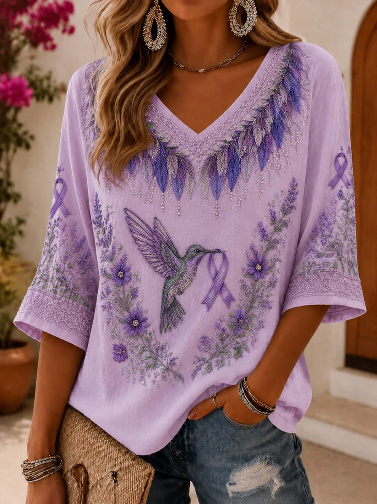 A woman wearing a lavender purple V-neck short-sleeve top with a large embroidered hummingbird and purple awareness ribbons, paired with distressed blue jeans.