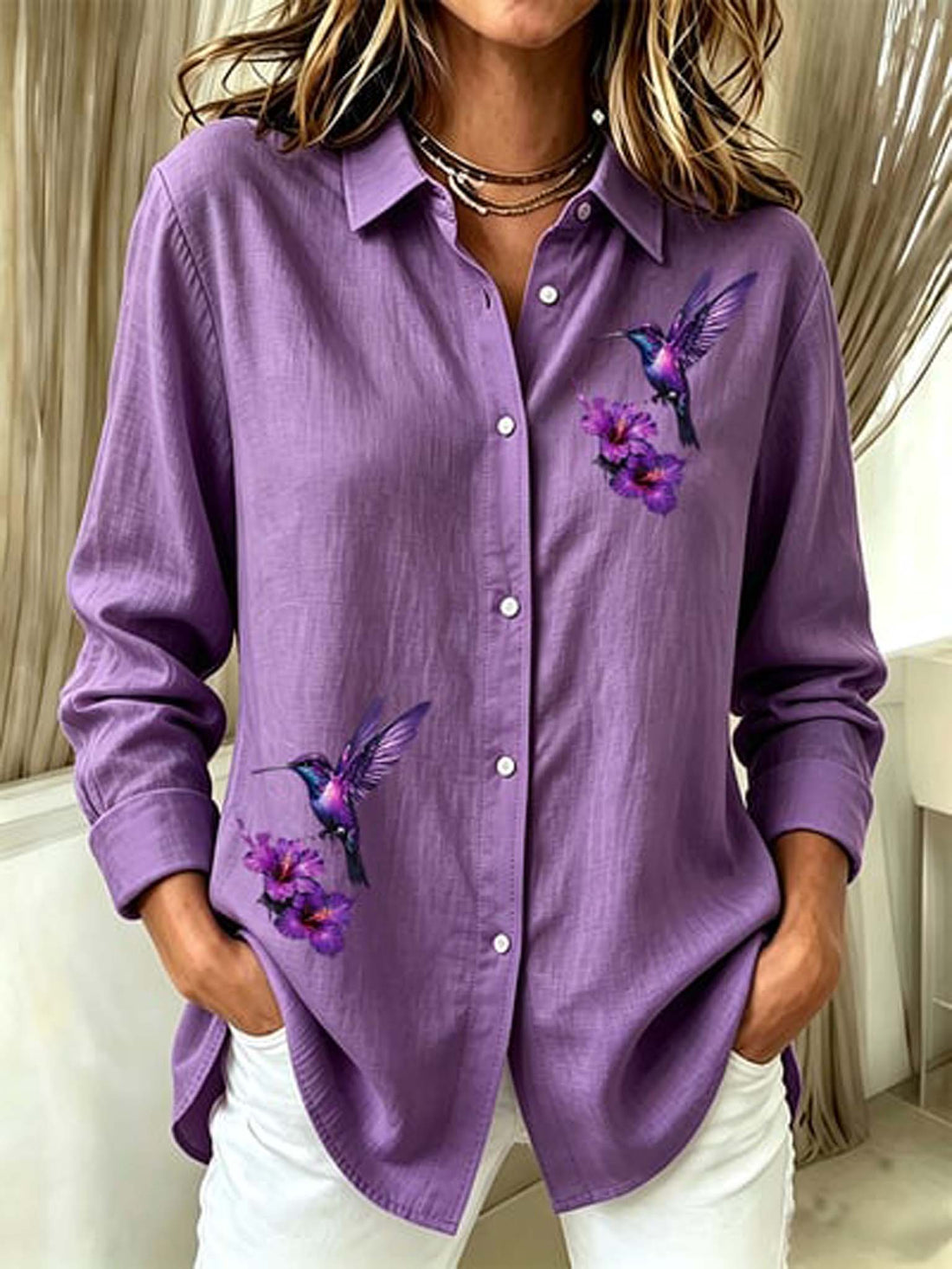 A woman wearing a lavender-purple long-sleeve button-up shirt with printed purple flowers and hummingbirds on the front, paired with white pants.
