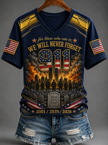 A navy blue V-neck graphic t-shirt with a detailed patriotic memorial design, hanging on a wooden hanger against a gray background. The shirt features '911' in a flag pattern, silhouettes of firefighters, tombstones, and American flags on the sleeves, paired with distressed denim shorts.