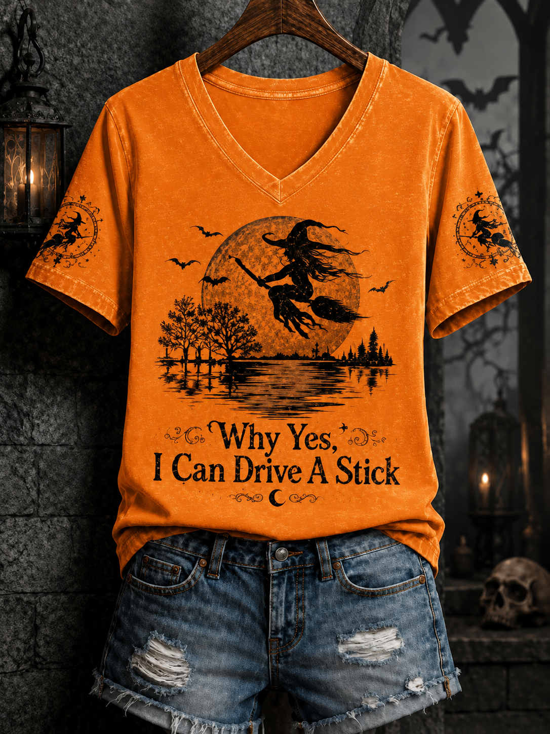An orange V-neck t-shirt with a Halloween graphic of a witch riding a broomstick in front of a full moon, displayed on a wooden hanger with distressed denim shorts against a dark gothic-themed background.