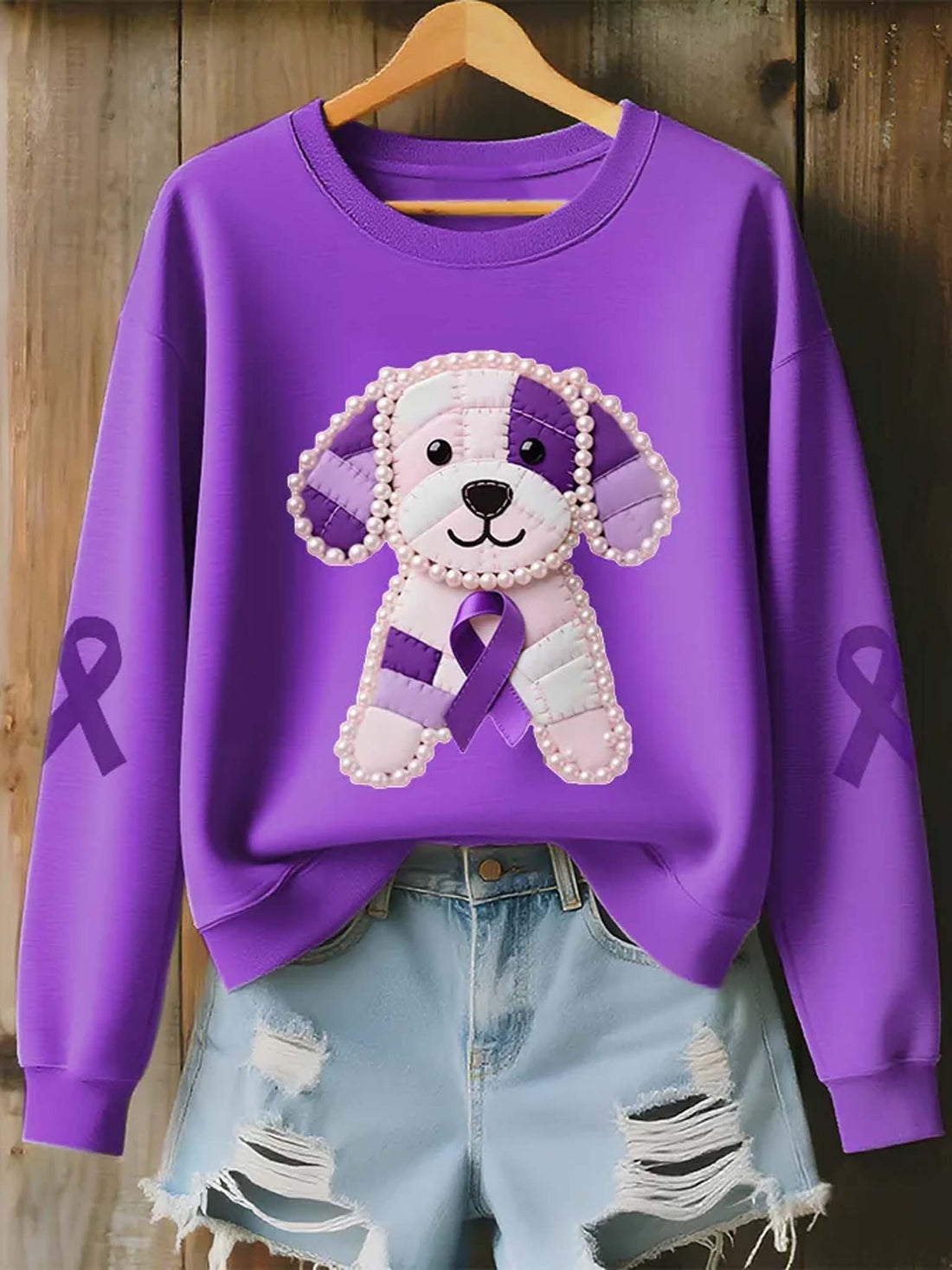 A purple crewneck sweatshirt with a dog patchwork design wearing a purple awareness ribbon, styled on a wooden hanger with light blue ripped denim shorts against a rustic wood plank background.