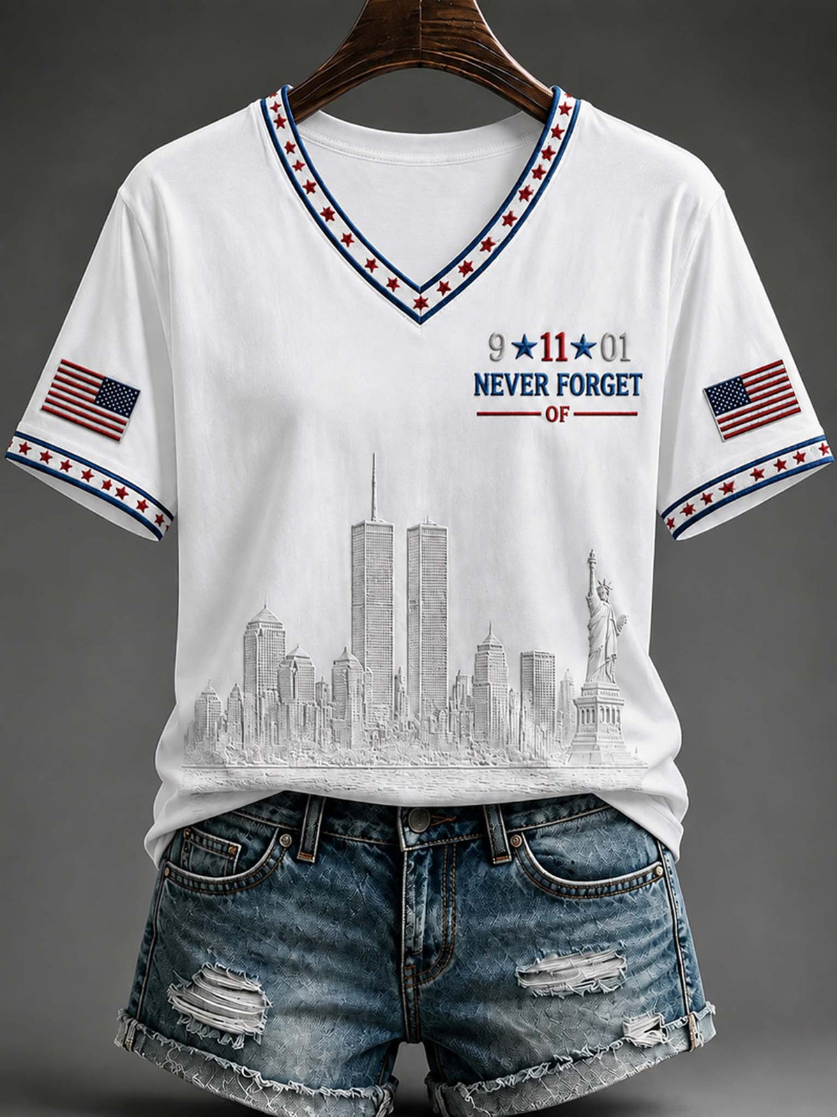 White V-neck t-shirt with a patriotic design featuring the New York City skyline, '9-11-01 NEVER FORGET' text, and American flag prints on the sleeves, styled on a hanger with distressed denim shorts against a dark gray background.