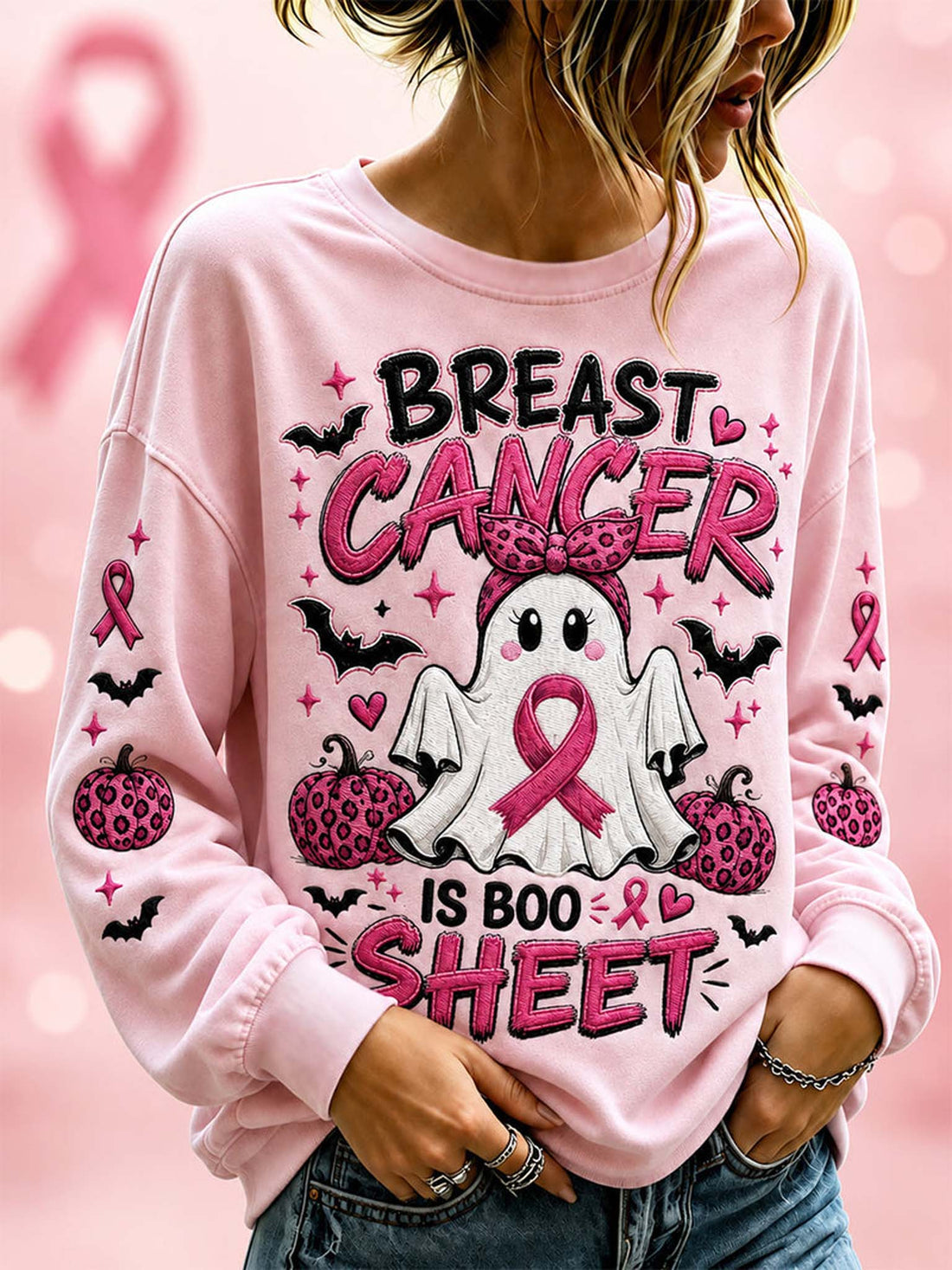A woman wearing a light pink long-sleeve sweatshirt with a 'Breast Cancer is Boo Sheet' Halloween graphic featuring a ghost, pumpkins, bats, and pink ribbons.